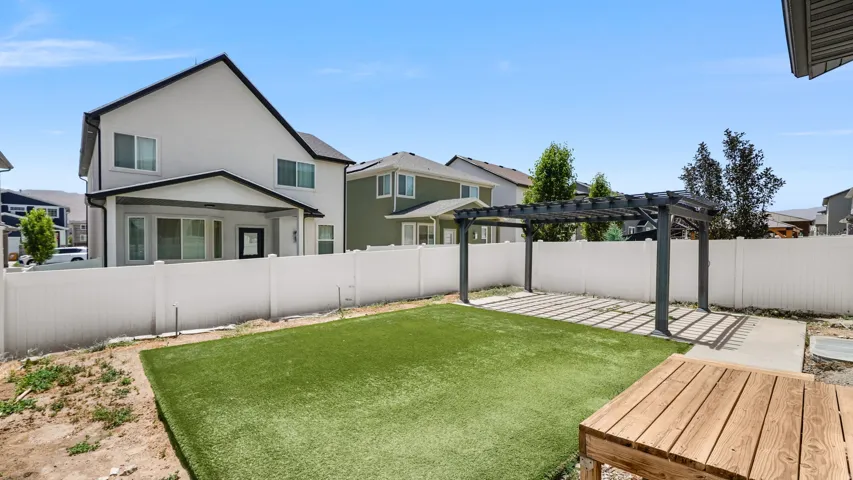 Spacious backyard featuring artificial turf, a white privacy fence, a dark-toned pergola over a concrete patio, and a wood-finish deck