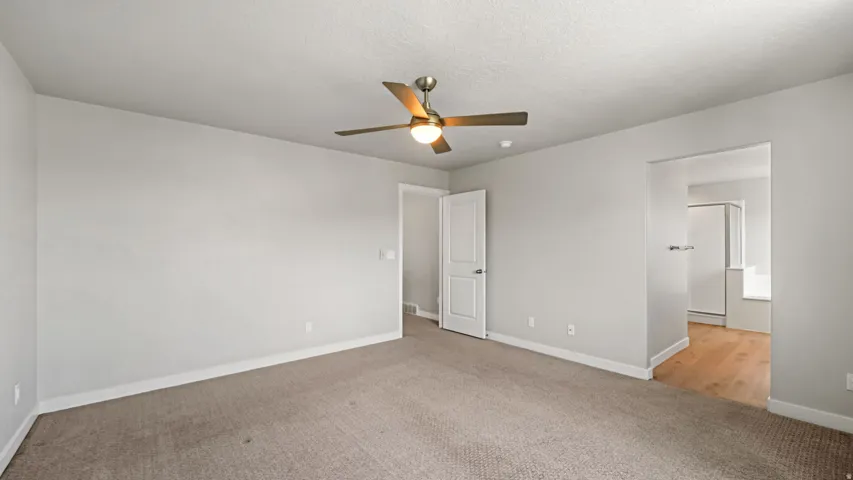 Spacious interior featuring neutral carpeting, light gray walls, a ceiling fan with integrated lighting, and an open doorway leading to a bathroom with wood-finish flooring and a built-in tub