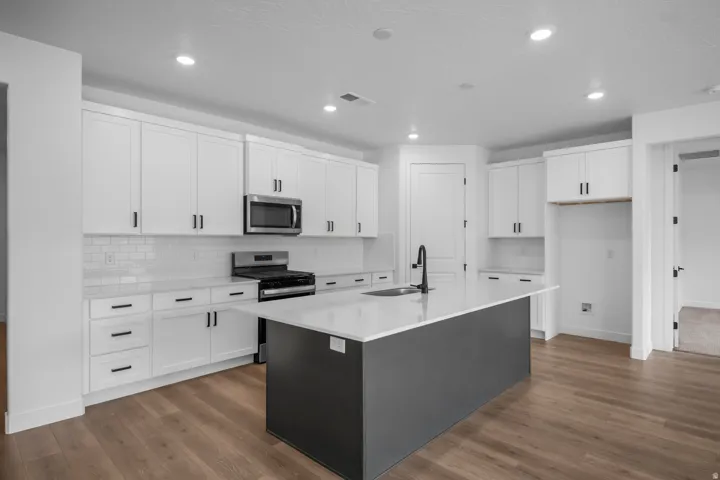 Modern kitchen featuring a central island with an integrated sink, white shaker cabinetry, white subway tile backsplash, stainless steel microwave, and wood-finish flooring