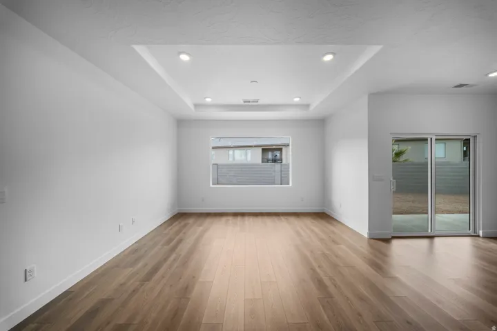 Spacious room featuring wood-finish flooring, a recessed tray ceiling with integrated lighting, a large window, and a sliding glass door