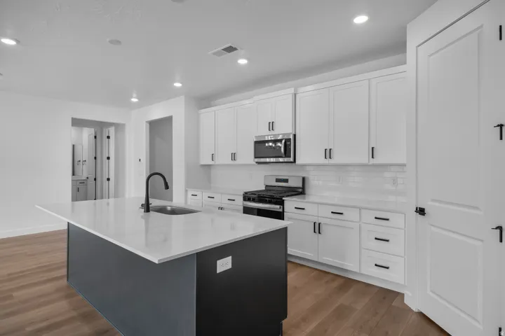 Modern kitchen featuring an expansive center island with an undermount sink and matte black faucet, wood-finish flooring, white shaker cabinetry, stainless steel appliances, and a subway tile backsplash