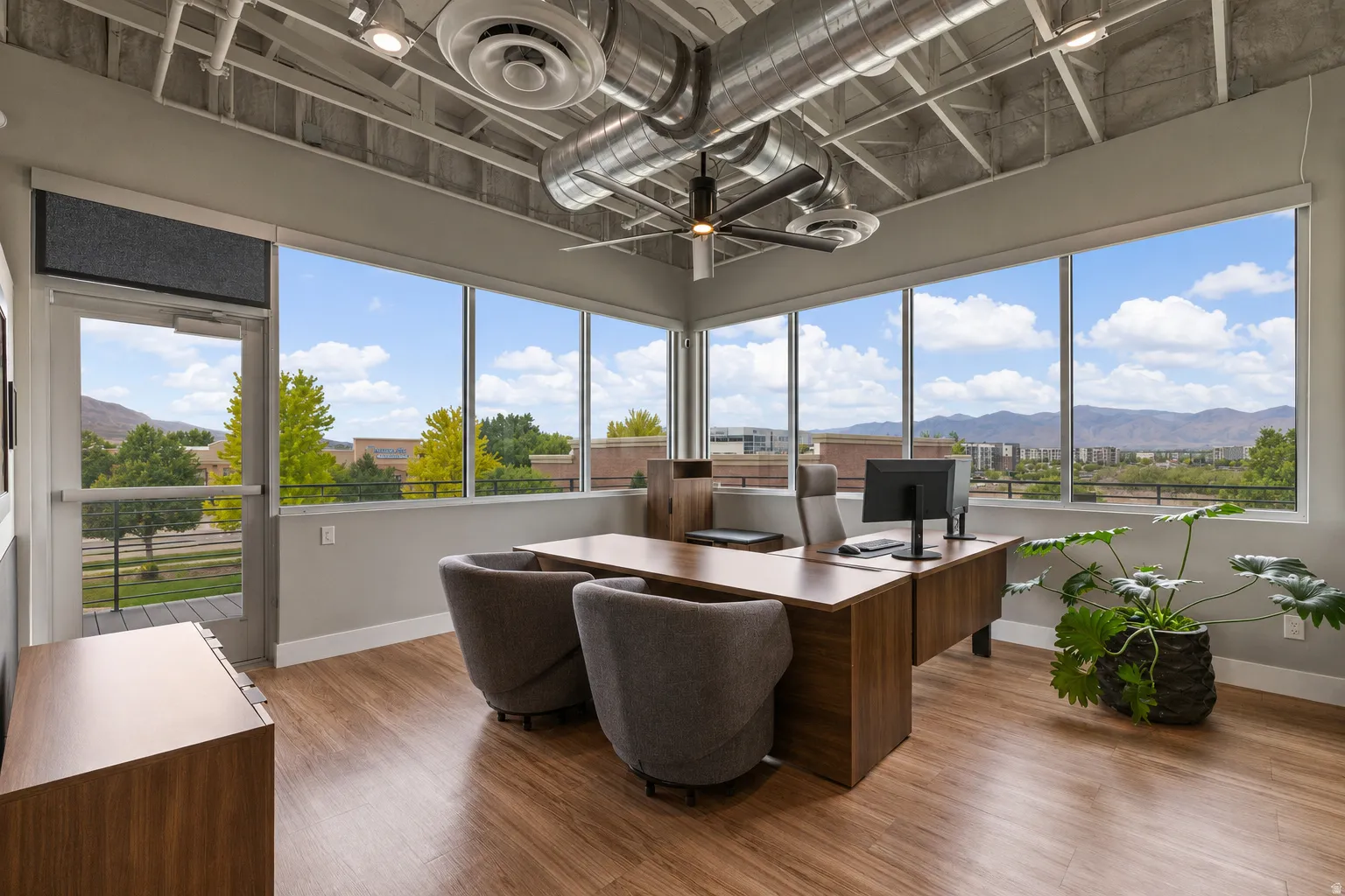 Corner office space featuring extensive window walls, exposed industrial-style ceiling, wood-finish flooring, a built-in wood-tone desk, and ceiling-mounted lighting with a fan