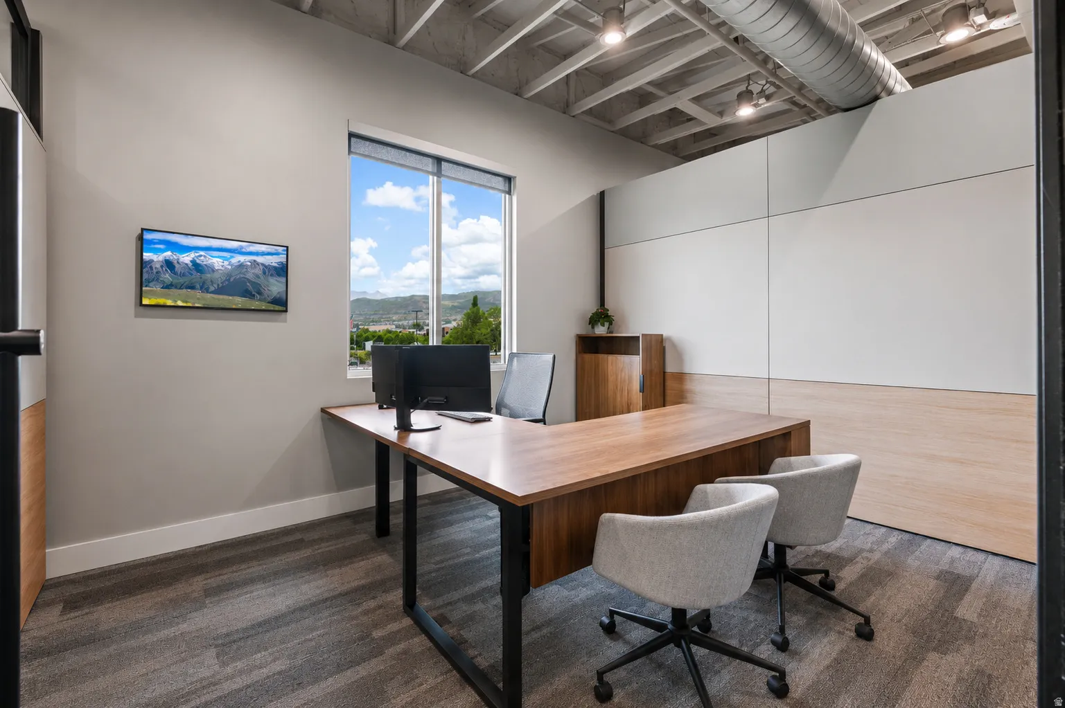 Contemporary office space featuring a large window with distant mountain scenery, wood-finish desk, exposed ceiling joists with track lighting, and carpeted flooring