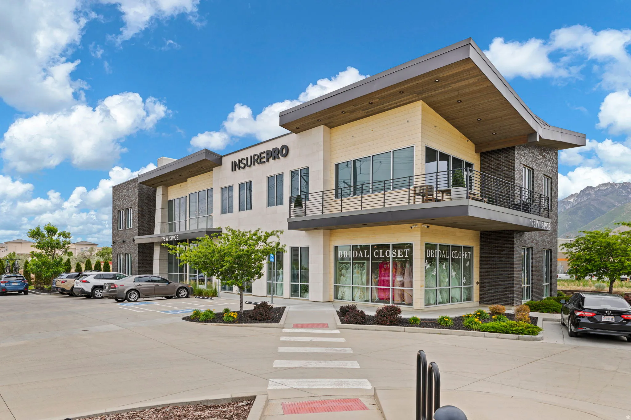Modern commercial building featuring stone and panel siding, large storefront windows, an upper-level balcony with metal railing, an angled roofline, and ample concrete parking