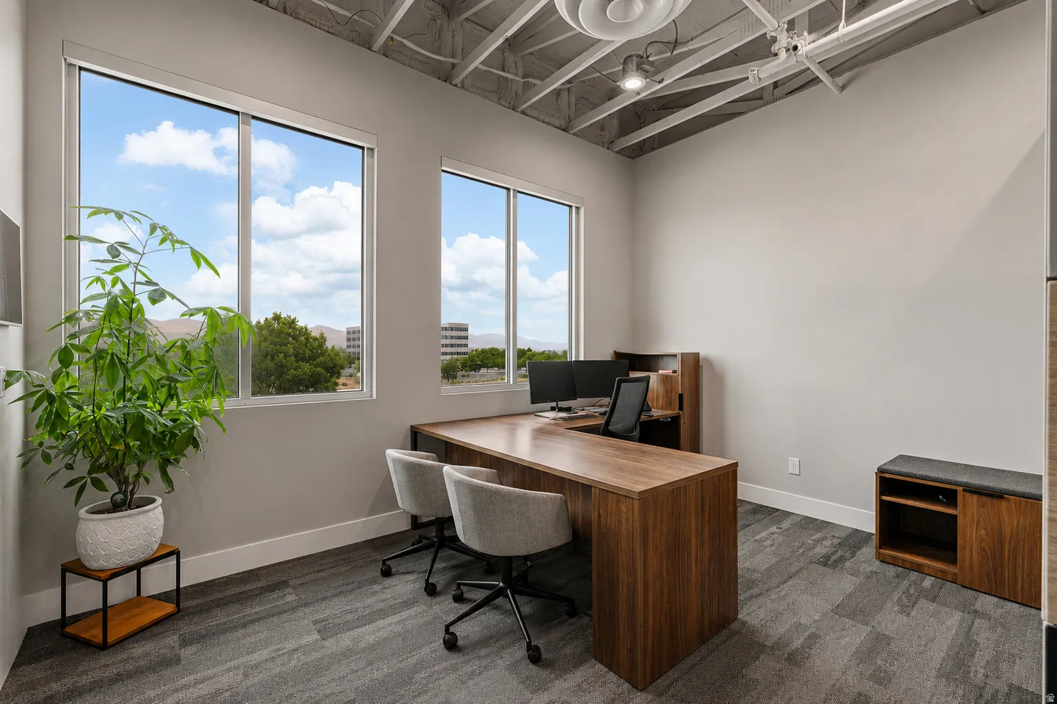 Modern office space featuring expansive windows, wood-finish desk, textured carpet, and an exposed ceiling
