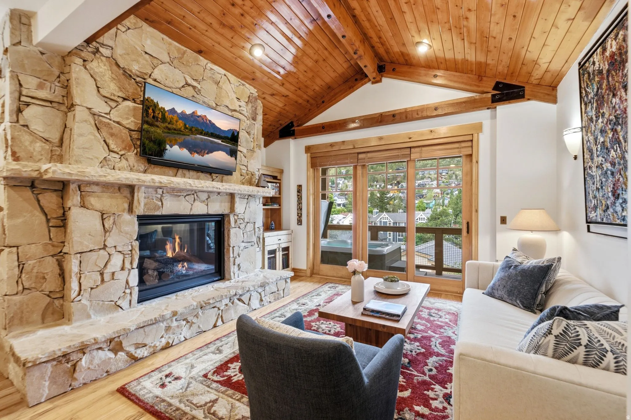 Vaulted wood plank ceilings with exposed beams, stone fireplace with hearth, built-in shelving, wood-finish flooring, and sliding glass doors opening to a balcony