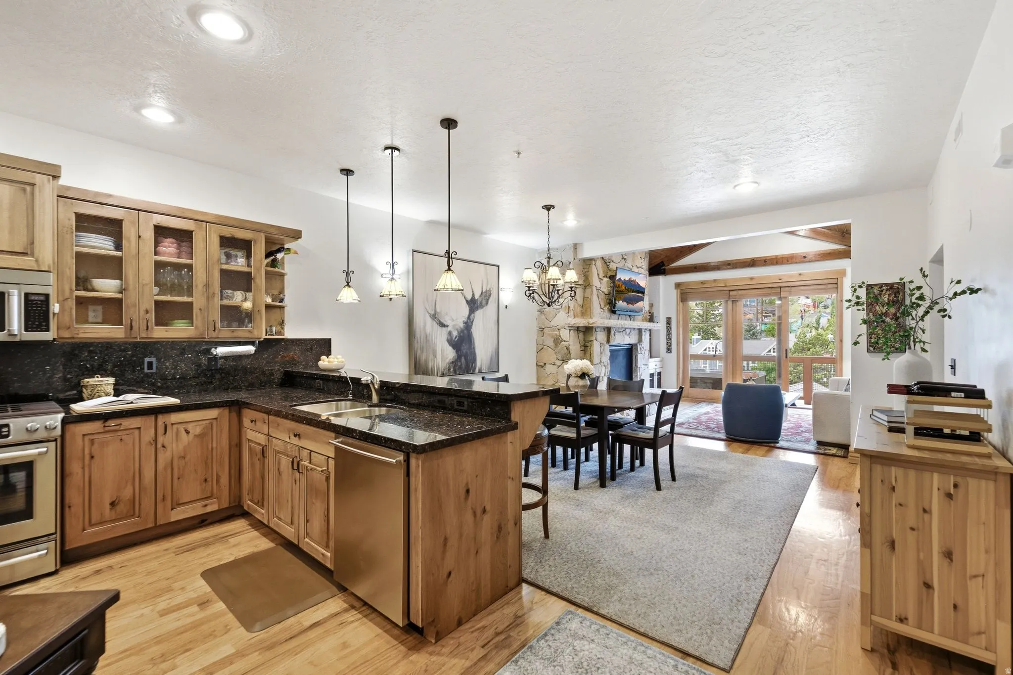 Open-concept kitchen and dining area featuring light wood cabinetry, dark stone countertops, stainless steel appliances, and wood-finish flooring