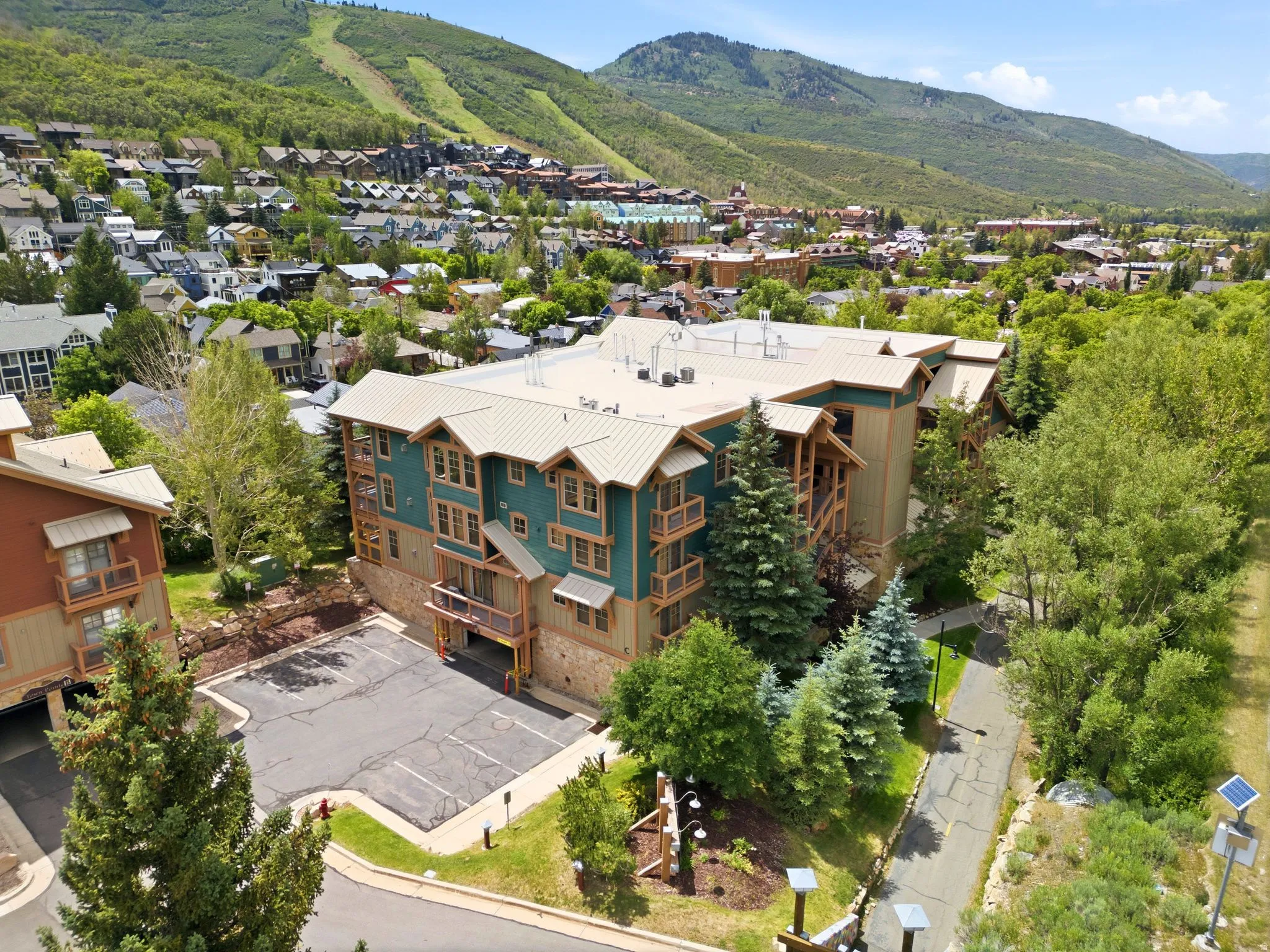 Multi-story building featuring a teal facade with wood accents and multiple balconies, a metal roof, and a paved parking area