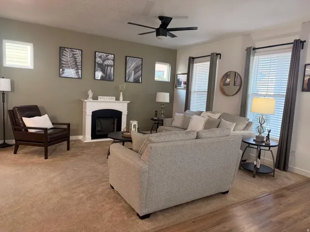 Living area featuring a white mantelpiece, dark wood-finish flooring, light tan carpeting, multiple windows with blinds, and a ceiling fan