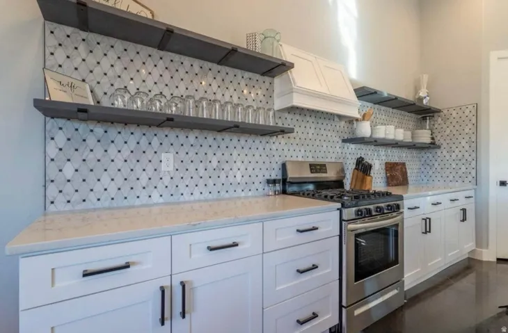 White shaker-style cabinetry with matte black hardware, stainless steel gas range, built-in range hood, light-colored countertops, and patterned tile backsplash