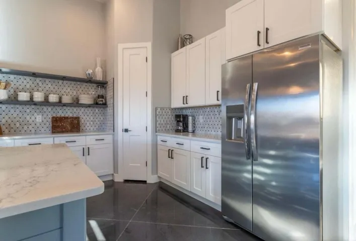 White shaker cabinetry with dark hardware, stainless steel refrigerator, patterned tile backsplash, open shelving, and a built-in pantry