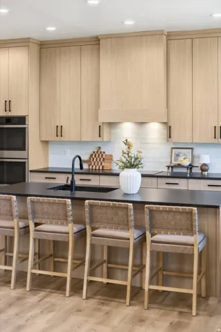 Kitchen featuring light wood-finish cabinetry with black hardware, a black countertop island with integrated sink, recessed ceiling lighting, a white subway tile backsplash, and built-in stainless steel double ovens