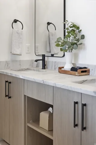 Dual vanity with a stone countertop, undermount sinks, wood-finish cabinetry, matte black hardware, and a framed mirror