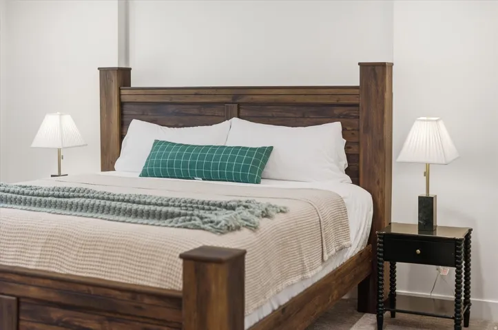 Spacious sleeping area featuring a robust wood-finish bed frame, light-colored walls, and a contrasting dark side table with a contemporary lamp