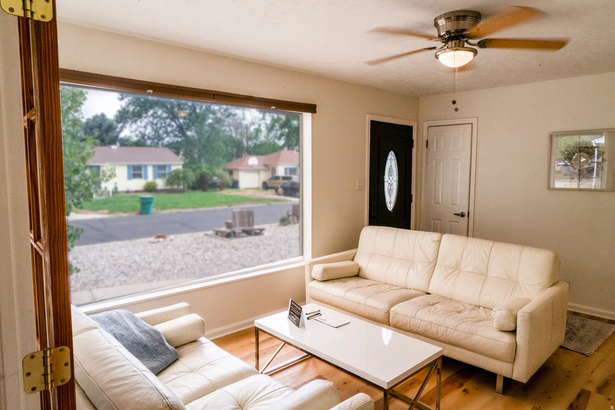 Living area featuring a large picture window, wood-finish flooring, a ceiling fan with light fixture, and a front entry door with an oval glass insert