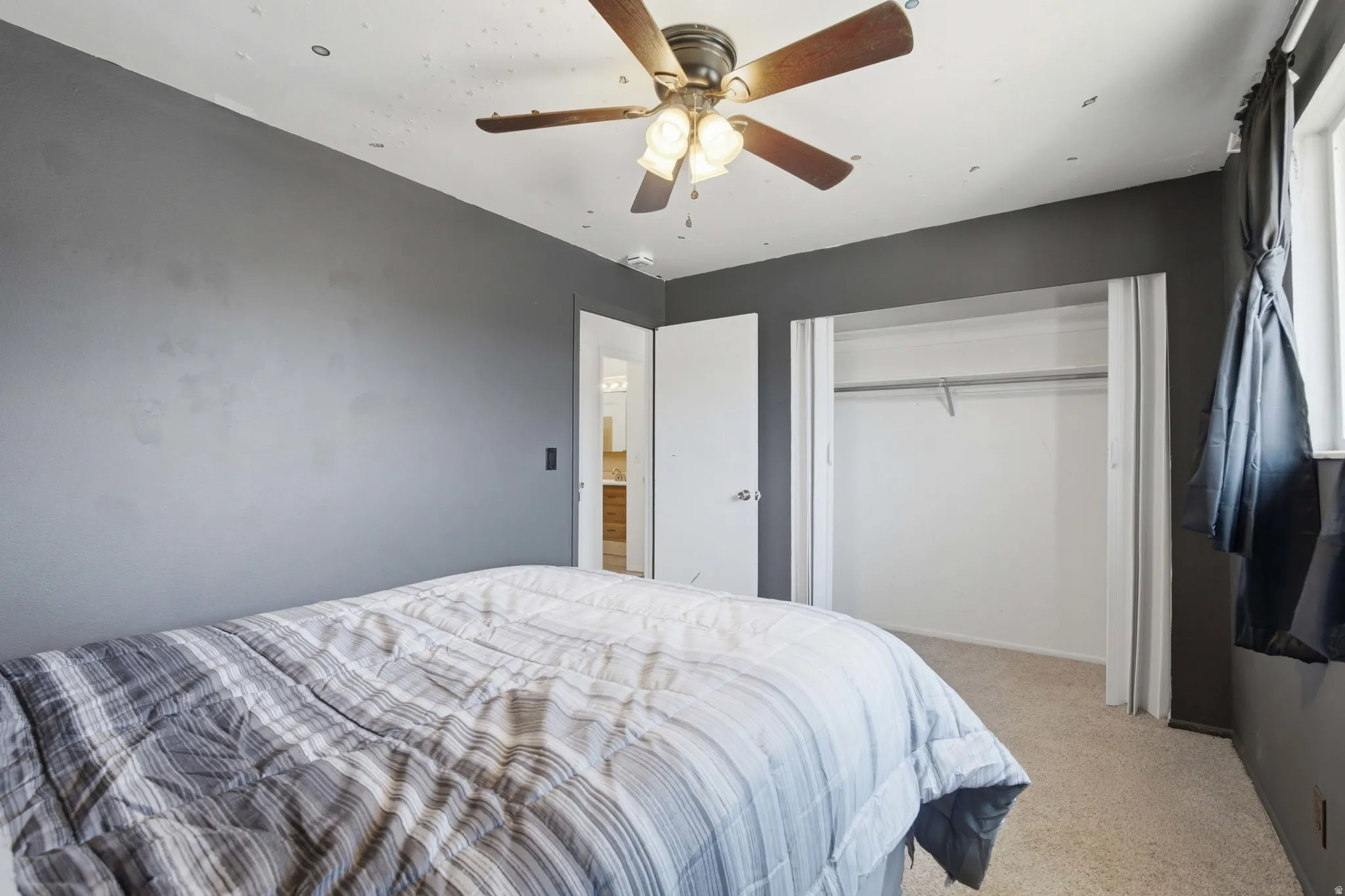 Carpeted interior space featuring a ceiling fan with integrated lighting, a built-in closet with bi-fold doors and clothing rod, and a window with roller shades