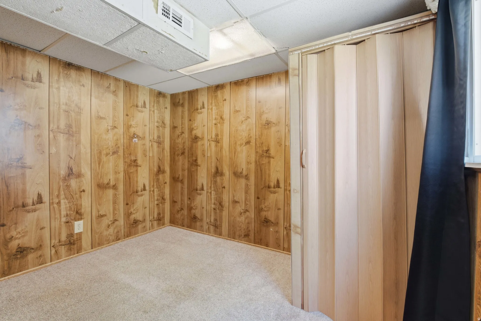 Carpeted room featuring wood paneling, drop ceiling, and a light-toned accordion door