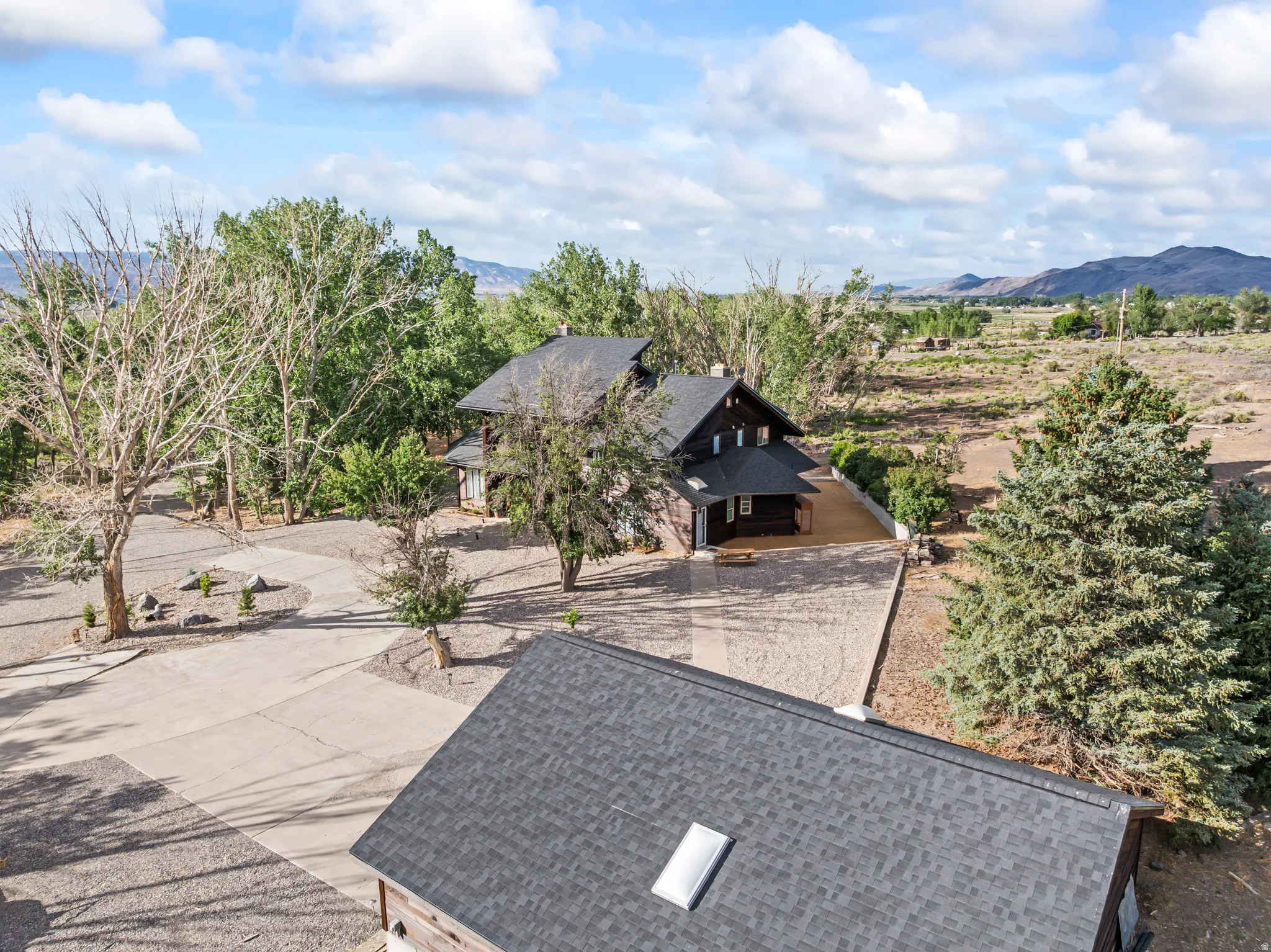 Expansive property with a dark-sided main residence, a concrete driveway, a gravel parking area, mature trees, and a mountain backdrop