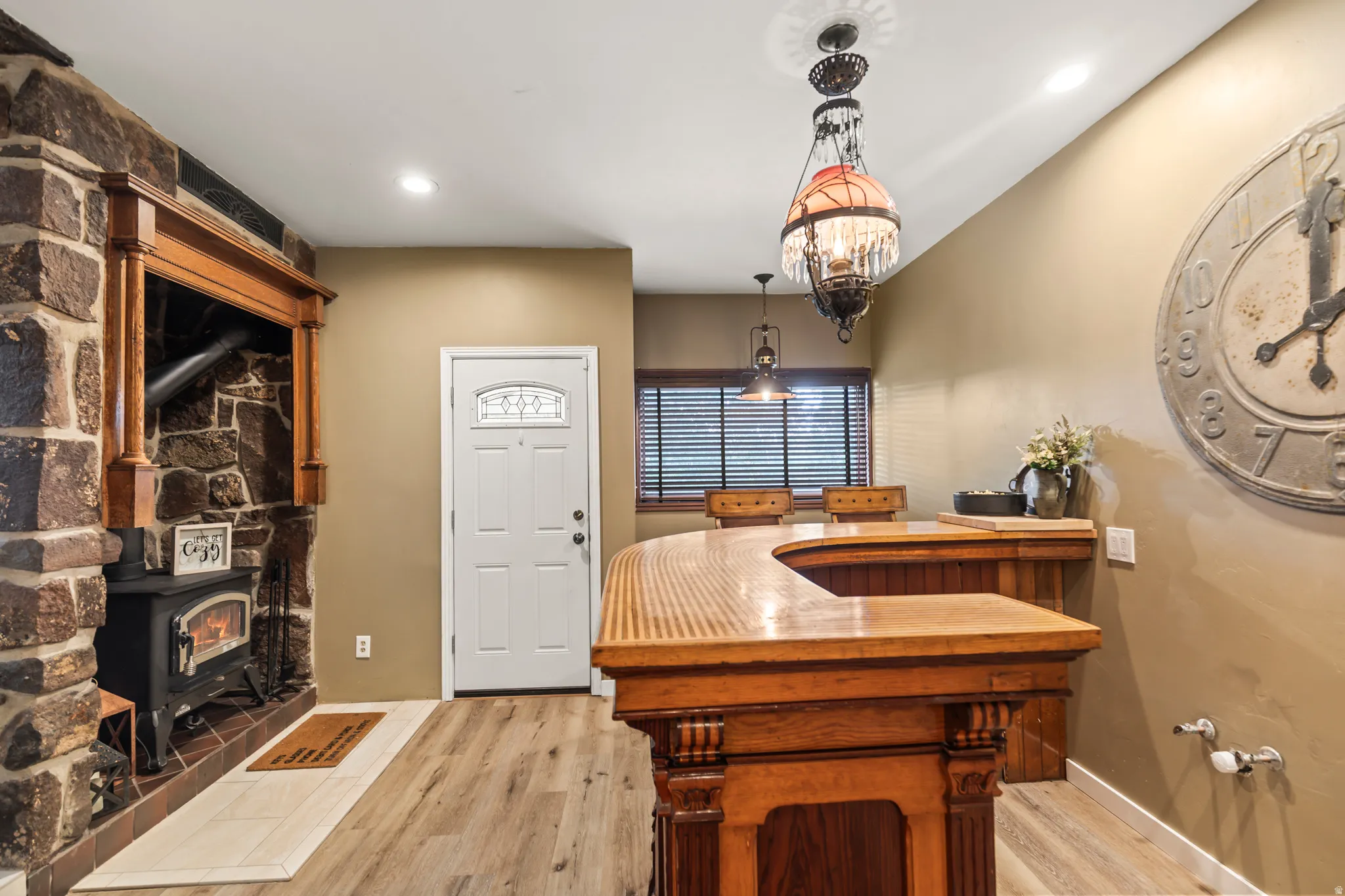 Stone fireplace with wood mantel, wood-burning stove, white entry door with decorative glass, wood-finish flooring, and a built-in wood bar