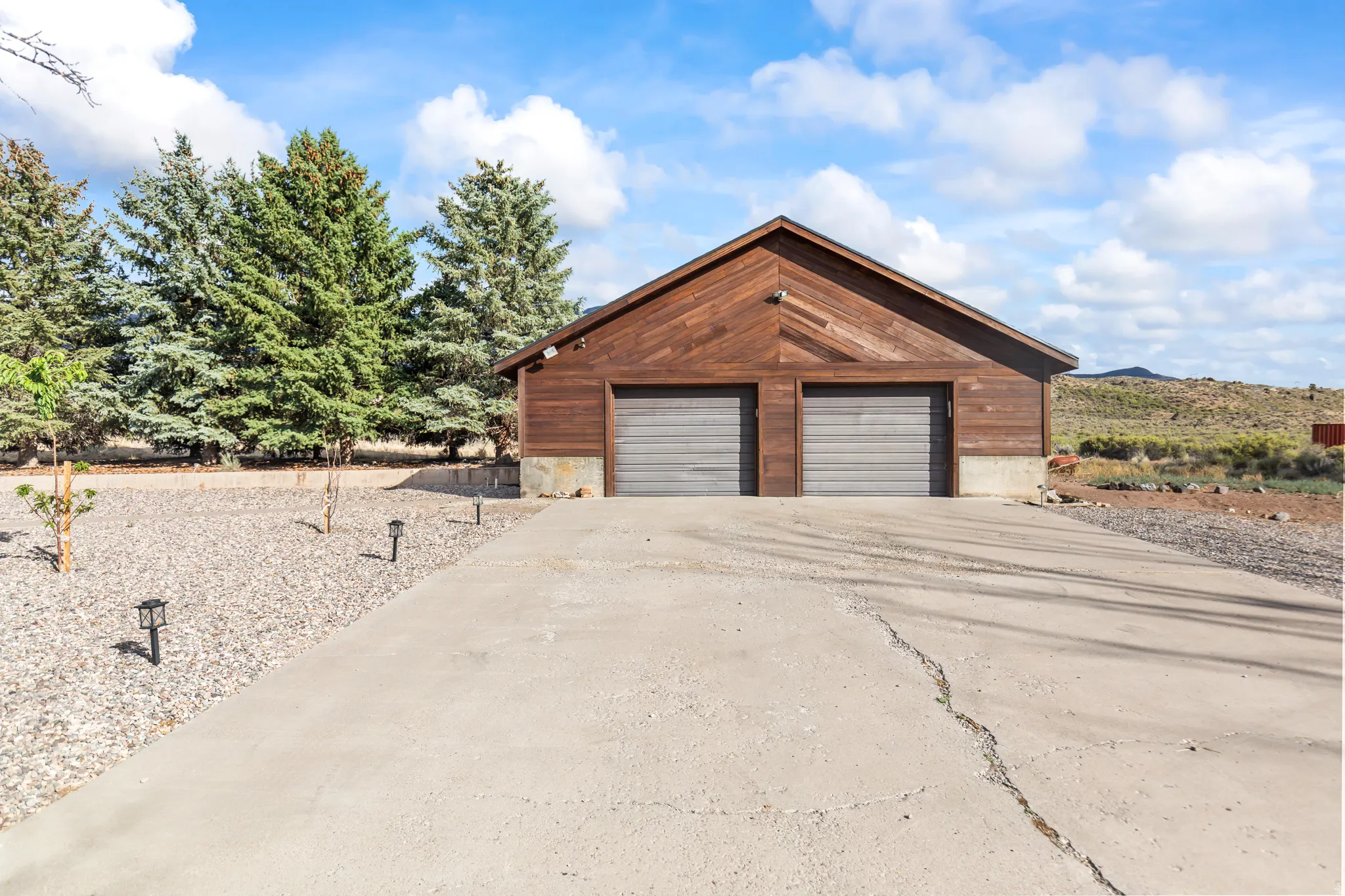 Wood-sided garage building featuring two roll-up garage doors, a concrete driveway, and gravel landscaping with mature evergreen trees