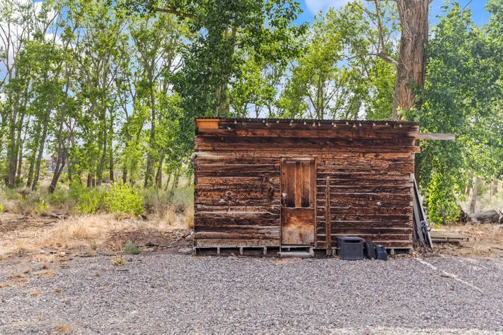 Rustic outbuilding with wood-plank siding and a wood door, surrounded by mature trees and a gravel ground cover