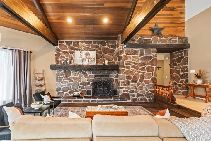 Spacious living area featuring a prominent stone fireplace, wood-finish vaulted ceiling with exposed beams, wood-finish flooring, and a stone accent wall