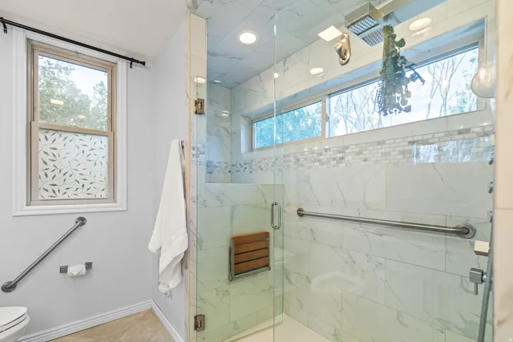 Frameless glass shower enclosure featuring marble-look tile, a mosaic accent band, a built-in wooden bench, and a rain shower head