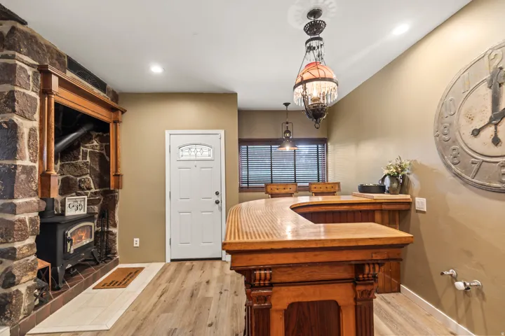 Stone fireplace with wood mantel, wood-burning stove, white entry door with decorative glass, wood-finish flooring, and a built-in wood bar