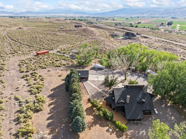 Expansive property featuring a multi-level residence with a dark shingle roof, surrounding mature trees, a detached accessory structure, a paved driveway, and natural, arid landscaping