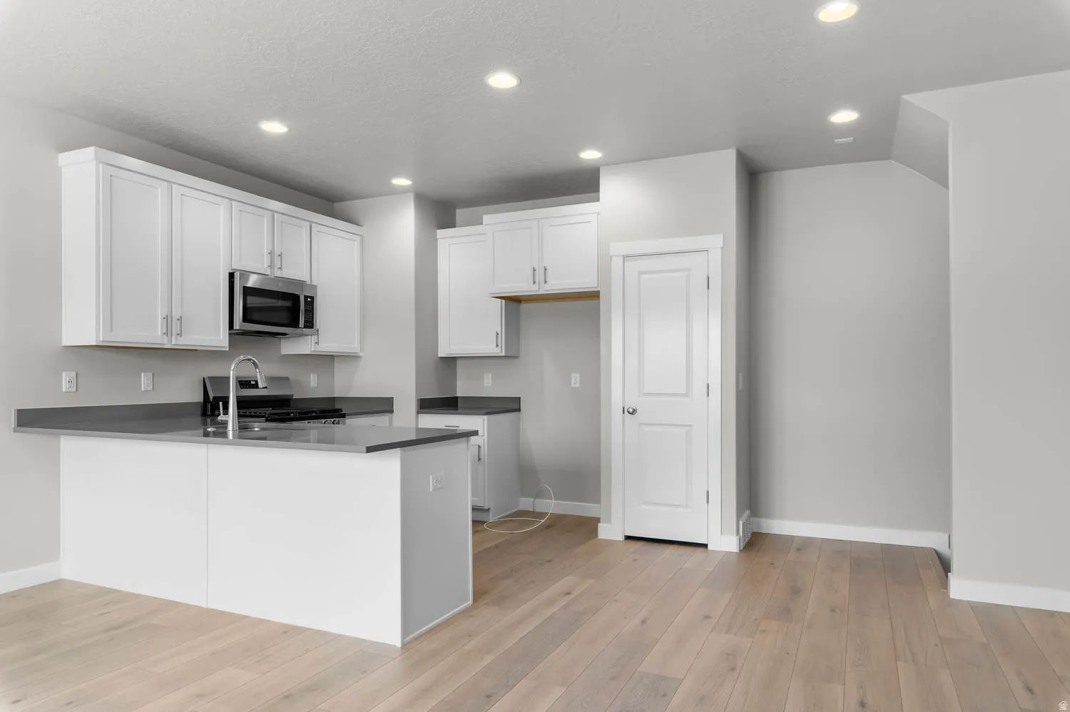 Contemporary kitchen featuring light wood-finish flooring, white cabinetry, dark countertops, a central island with an integrated sink, and stainless steel appliances including a built-in microwave