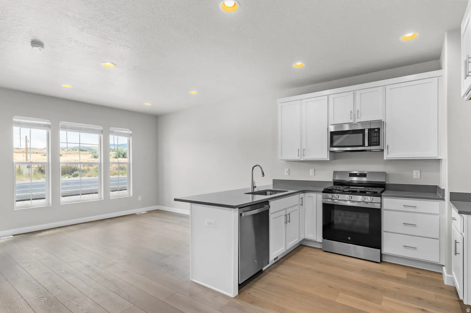 Modern kitchen featuring white cabinetry, stainless steel appliances, dark countertops, a central island with a sink, and wood-finish flooring extending into the open living space