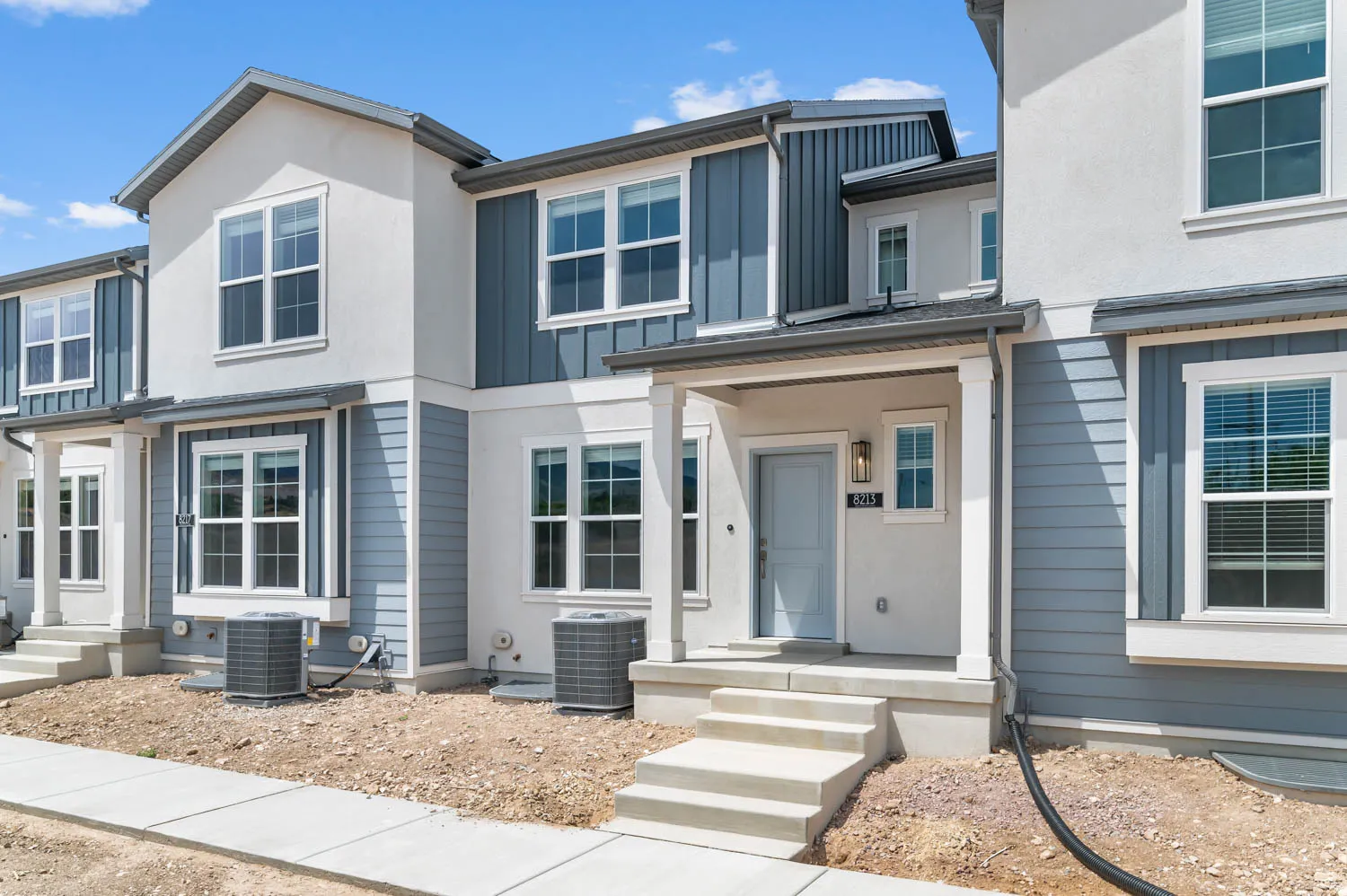 Contemporary townhouse exterior featuring a covered porch entry, multi-pane windows, vertical board and batten siding, horizontal lap siding, and a light-colored stucco finish