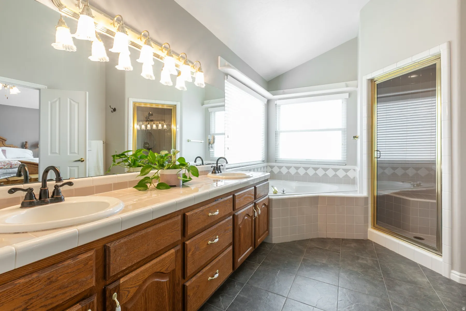 Spacious bathroom featuring a dual vanity with a tile countertop, a large soaking tub with tile surround, a separate shower enclosure with a gold-toned frame, and dark tile flooring