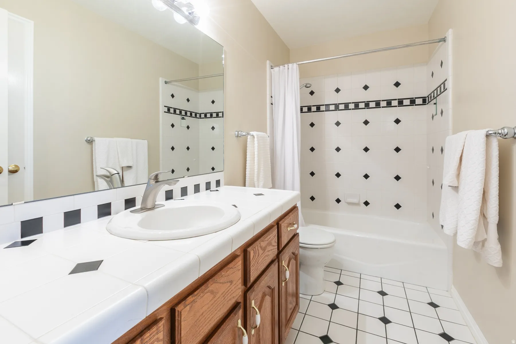 Bathroom featuring a white tile vanity with integrated sink, wood-finish cabinetry, full-width mirror, and a tub-shower combination with black and white accent tiling