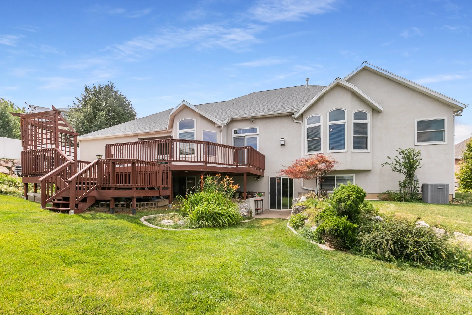 Expansive backyard featuring a multi-level wood deck with pergola, stucco exterior, mature landscaping, and sliding glass doors