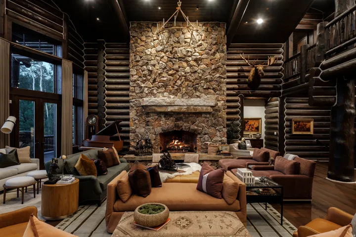 Great room featuring a floor-to-ceiling stone fireplace, exposed log walls, dark wood-finish flooring, a two-story window wall, and an upper-level balcony