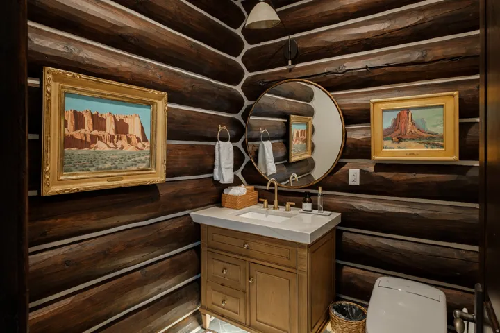 Powder room featuring log walls, a wood vanity with a white countertop, a circular mirror, and brass fixtures