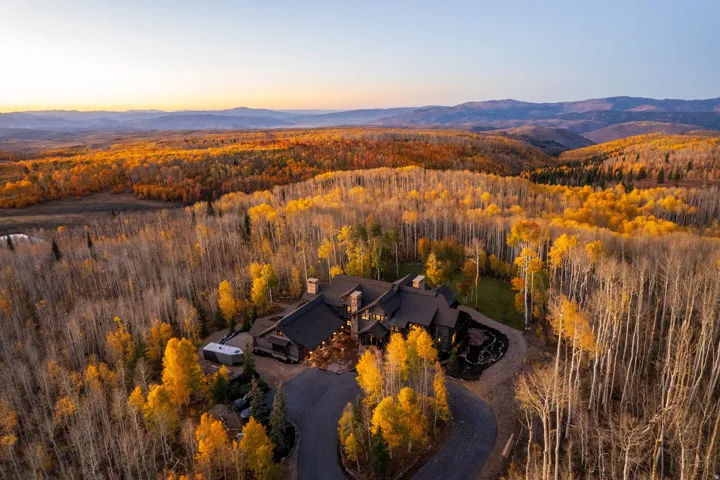 Mountain estate with a winding paved driveway, surrounded by dense deciduous trees and distant mountain ranges
