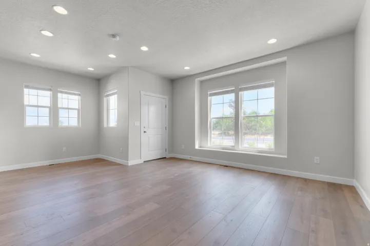 Spacious interior room featuring wood-finish flooring, ample natural light from multiple windows, recessed ceiling lighting, and a white paneled door