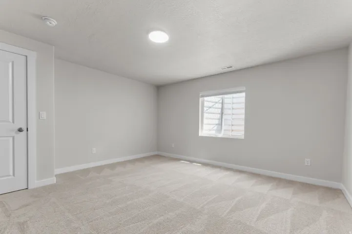 Spacious room featuring light-toned walls, neutral carpeting, a single window with blinds, a recessed ceiling light, and a white interior door