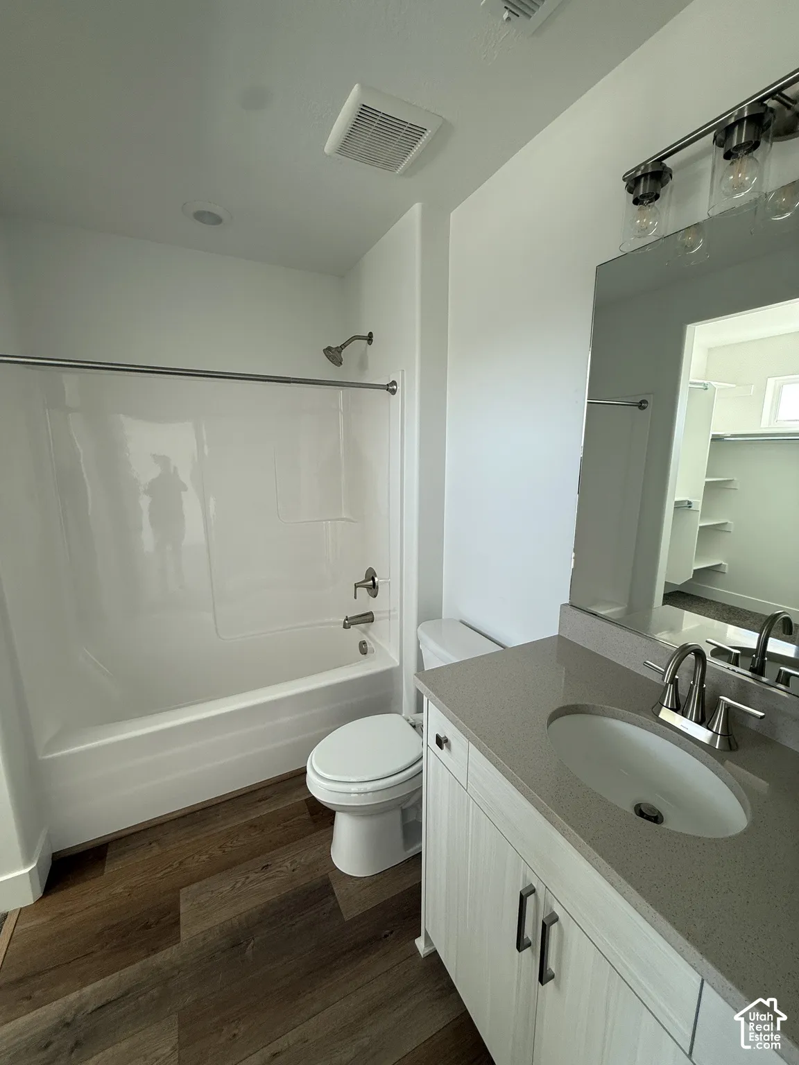 Wood-finish flooring, white bathtub with shower, toilet, vanity with integrated sink, and wall-mounted light fixture