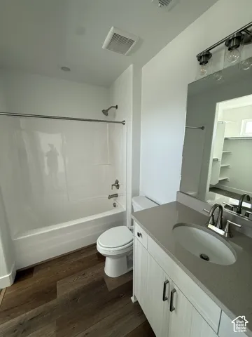 Wood-finish flooring, white bathtub with shower, toilet, vanity with integrated sink, and wall-mounted light fixture
