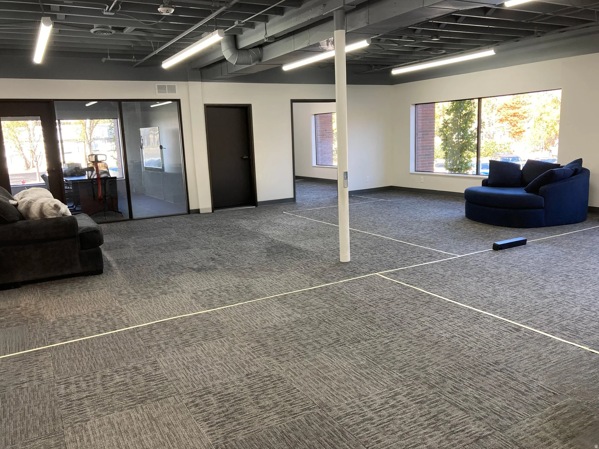 Open concept space featuring modular carpet tiles, extensive natural light from large windows, exposed ceiling with ductwork, and fluorescent lighting