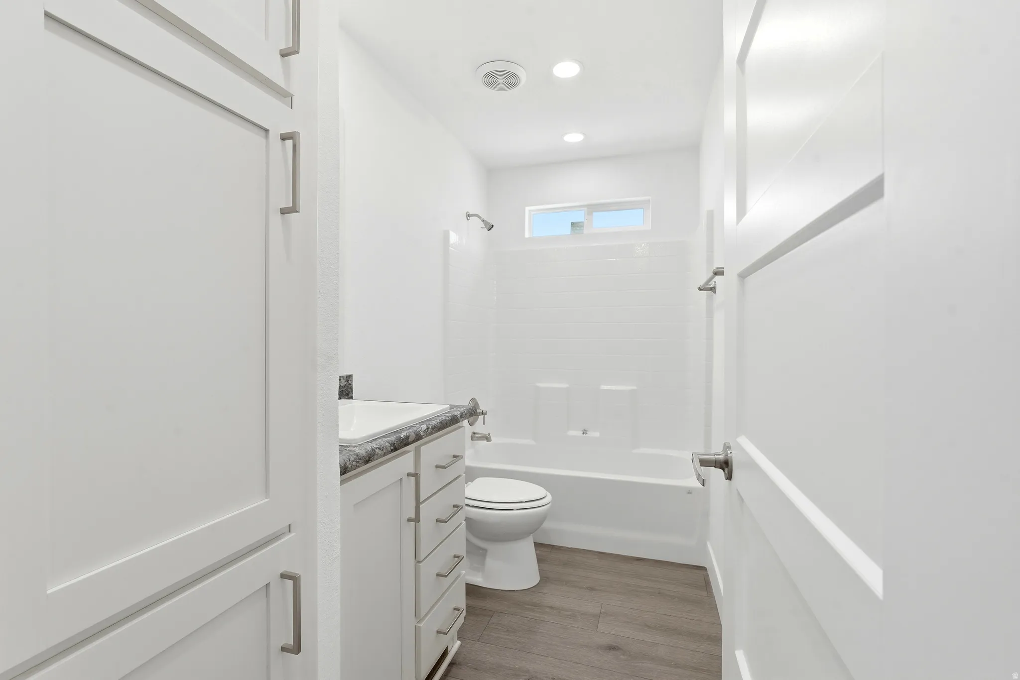 Full bathroom with shaker-style cabinetry, a vanity with a dark patterned countertop, wood-finish flooring, a white tub and shower combination, and a horizontal window