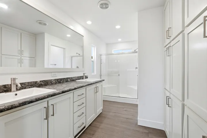 Bathroom featuring a dual vanity with undermount sinks, a large wall-mounted mirror, extensive white cabinetry, a glass-enclosed shower, and wood-finish flooring