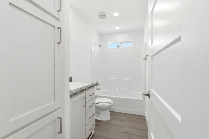 Full bathroom with shaker-style cabinetry, a vanity with a dark patterned countertop, wood-finish flooring, a white tub and shower combination, and a horizontal window