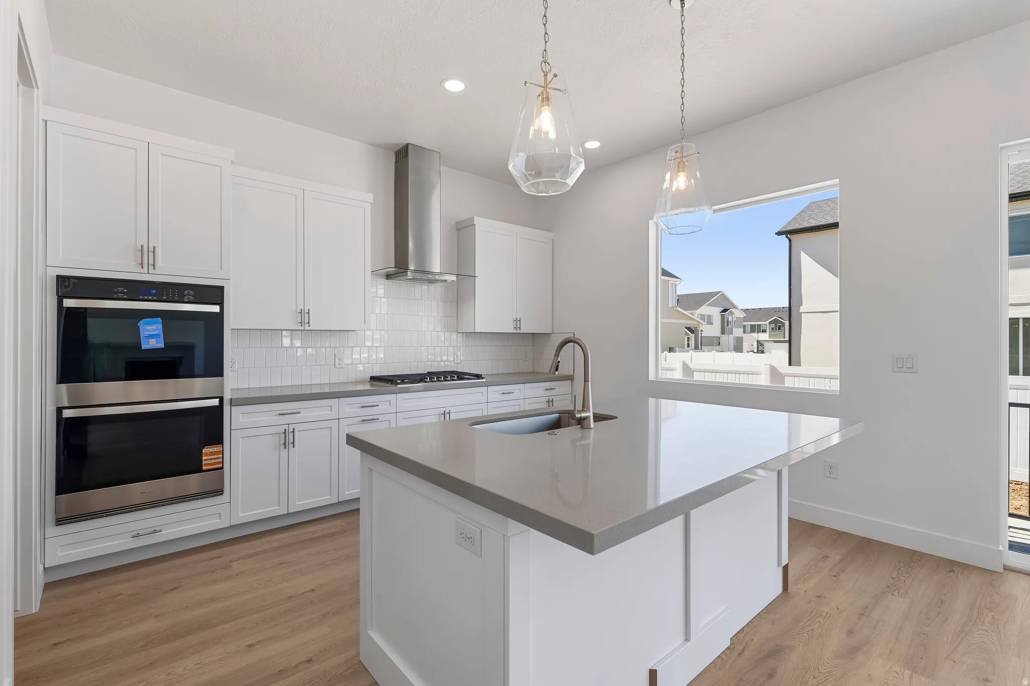 Bright kitchen featuring white cabinetry, light-toned countertops, stainless steel double ovens, a matching range hood, and wood-finish flooring