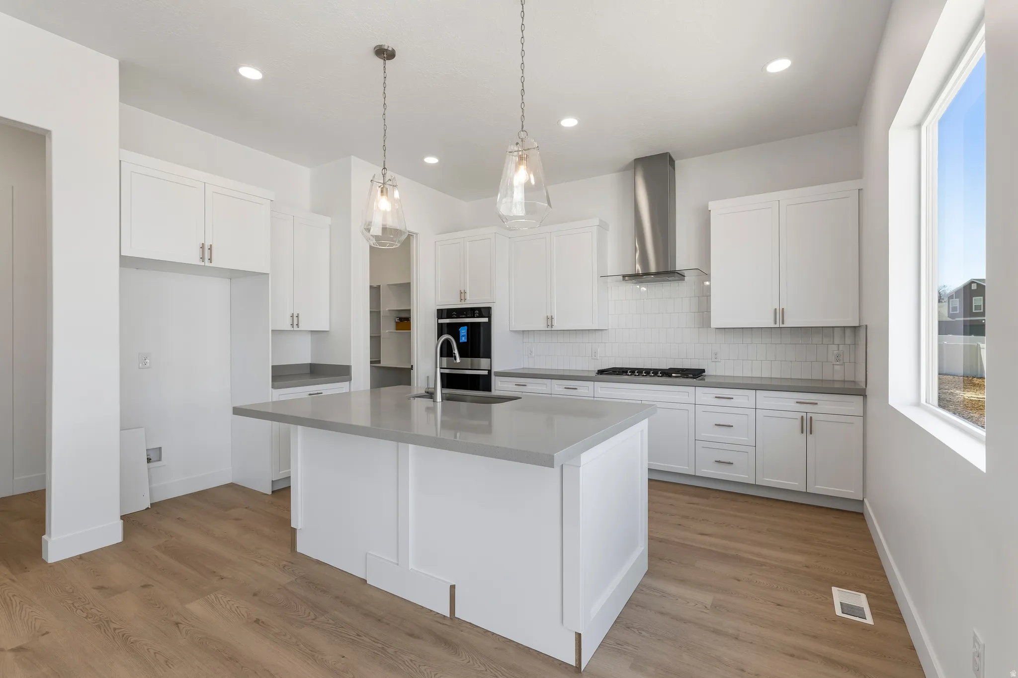Modern kitchen featuring a central island with a sink, white shaker cabinetry, light-toned countertops, wood-finish flooring, and pendant lighting