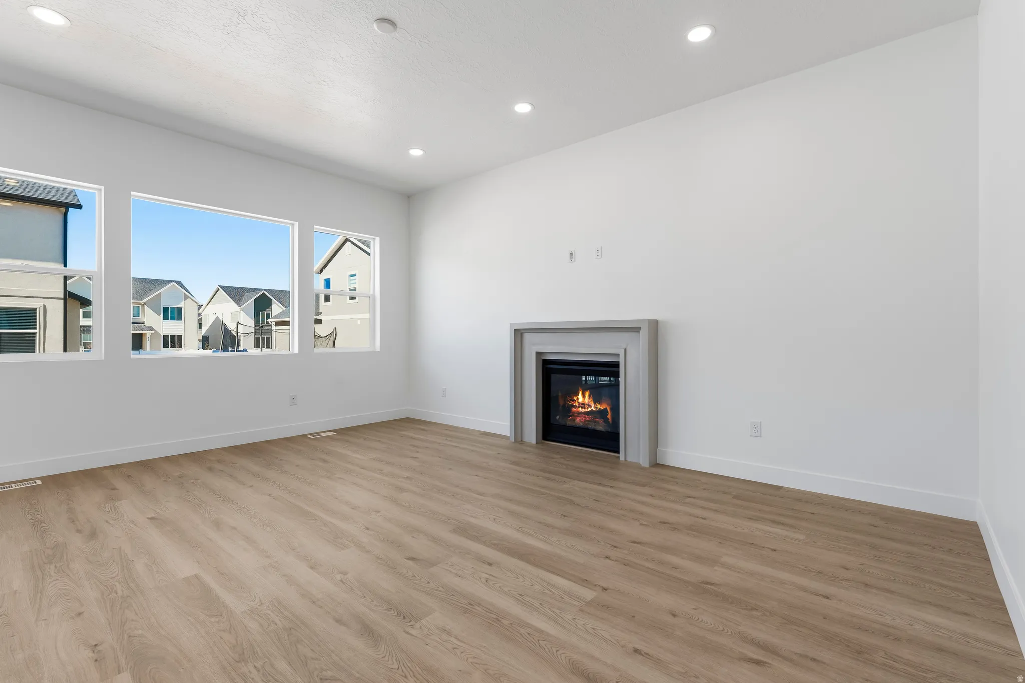 Spacious room featuring wood-finish flooring, a contemporary fireplace with a light gray surround, multiple large windows, recessed lighting, and white baseboards