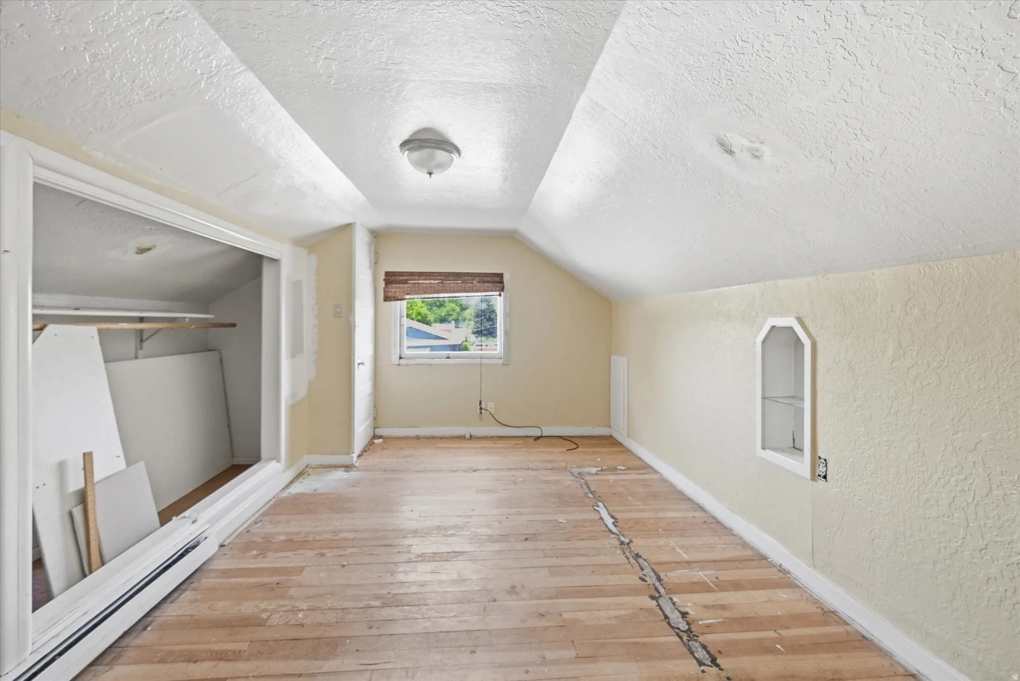 Attic room featuring sloped ceilings, exposed wood-finish flooring, a single window with blinds, and a built-in wall niche
